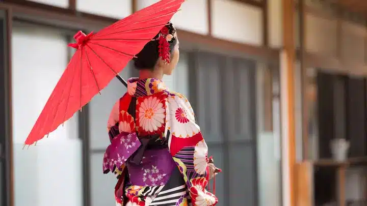 Woman in a colorful floral kimono seen from behind, holding a red parasol outside a traditional building area.