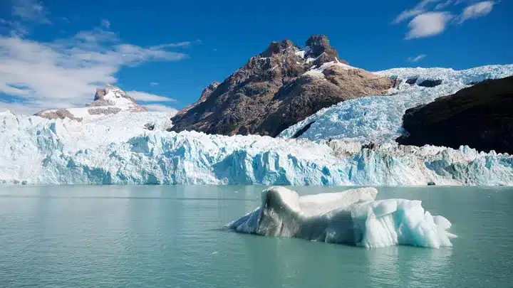 Glacier-fed lake with a blue iceberg floating in calm water and rugged snow-capped mountains in the background under a clear blue sky