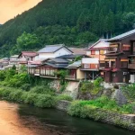 Traditional Japanese houses line a riverbank with lush green hills in the background at sunset, creating a peaceful riverside village scene.