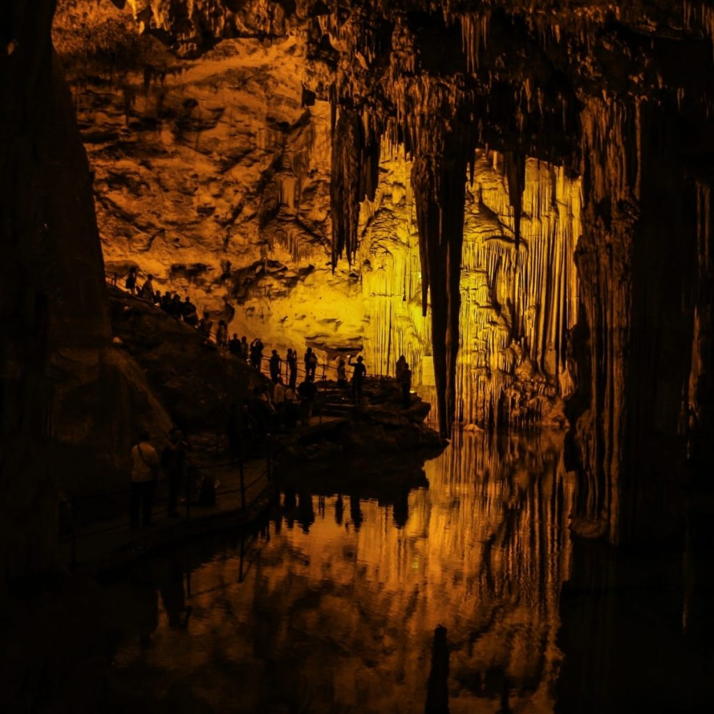 Inside a cavern with towering stalactites and amber lighting; groups of people walk along a guarded path beside a reflective pool.