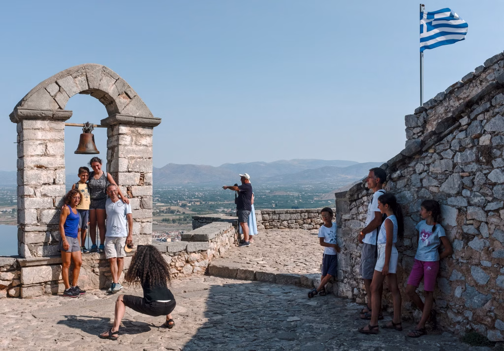Visitors posing on a stone battlement beside a bell arch, with a Greek flag and a bay below.