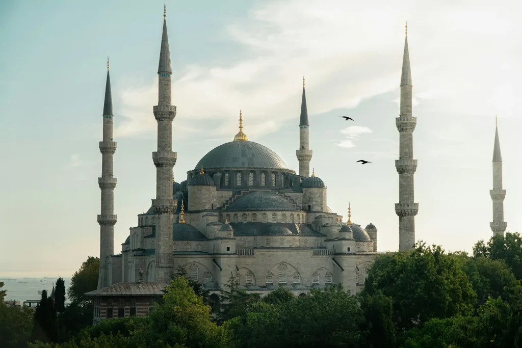 Grand mosque with multiple minarets and a large central dome, surrounded by greenery, with the sea in the background.