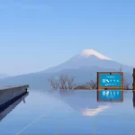 Infinity pool with a calm blue surface reflecting Mt. Fuji under a clear sky; a wooden railing and blue sign are visible in the background.