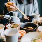 A person uses chopsticks to pick food from a bowl at a table filled with multiple bowls of Japanese-style dishes.