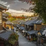 Historic street of traditional Japanese machiya houses at sunset; tiled roofs, wooden storefronts, and satellite dishes visible on the right.
