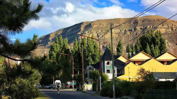 A sunlit residential street with bright yellow and green houses, trees, and a cyclist riding toward the camera, with a rocky hill in the background.