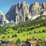 Green valley with scattered houses and pine trees at the foot of rugged snowy mountains under a clear blue sky.
