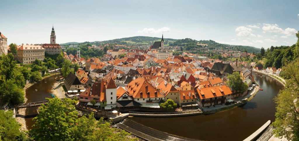 Panoramic view of a historic town with red-tiled roofs along a winding river and green hills in the distance.