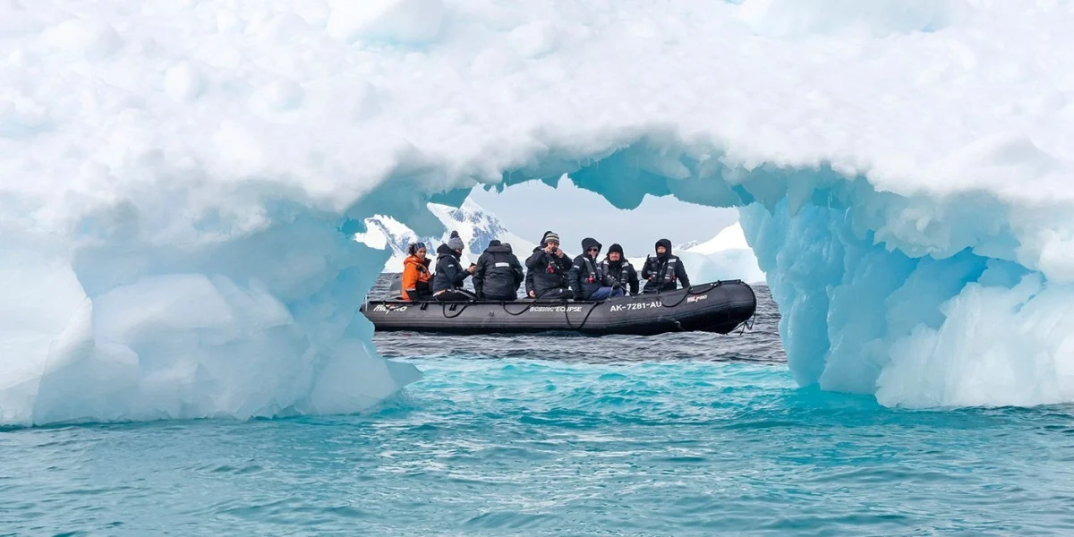 A small inflatable boat carries people in winter gear through a blue ice arch in Arctic waters.
