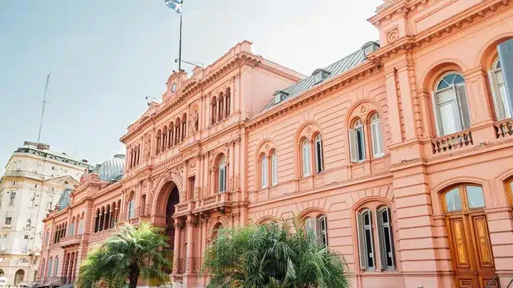 Pink neoclassical building with arched windows and palm trees in front, sunlit street scene
