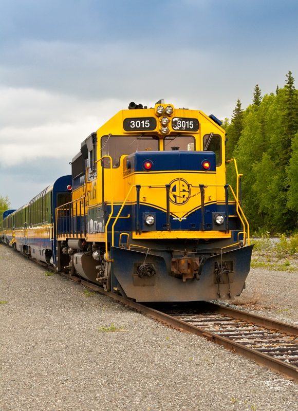 Front view of a bright yellow and blue diesel locomotive with the number 3015, on a gravel track with green trees nearby