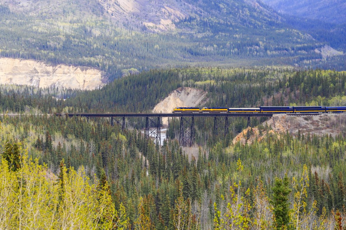 A blue-and-yellow passenger train crosses a tall iron bridge over a forested valley with mountains in the background.