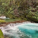 Cascading turquoise river rapids running through green jungle beside a wooden lookout railing.