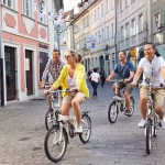 Group of four friends riding bicycles down a cobblestone street lined with colorful buildings on a sunny day.