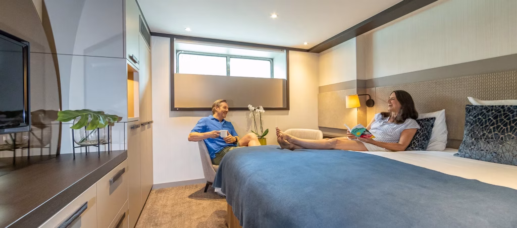 A couple relaxes in a modern hotel room: man in a blue shirt sits in a chair sipping coffee while a woman lies on the bed reading a magazine, with blue bedding and warm lighting.