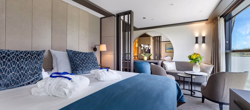 Luxurious hotel room with a large bed, blue-patterned pillows, and white towels on a blue bedspread; seating area and floral arrangement nearby, view through floor-to-ceiling windows.