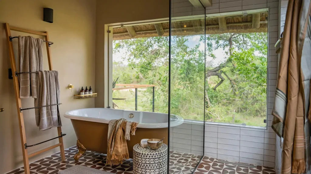 Bathroom with a freestanding tub beside a glass shower, and a wooden towel ladder in front of a beige wall; view of greenery outside the window.