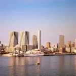 City skyline along a harbor with a large white cruise ship docked at the waterfront, glassy water reflecting the buildings at sunset.