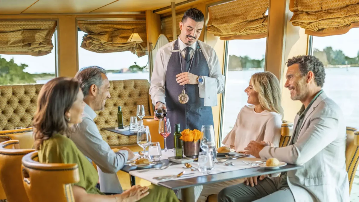 Waiter pours wine for a group of adults at a dining table inside a vintage train car, with smiles and wine glasses.