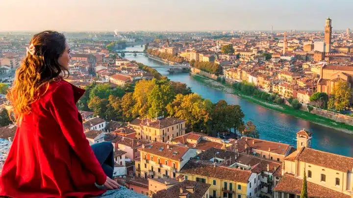 Woman in a red coat sits on a ledge overlooking a river through a historic town with terracotta roofs and lush green trees A scenic overlook.