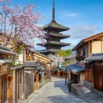 Traditional Japanese street with wooden houses, a stone-paved path, blooming cherry blossoms, and a multi-tier pagoda in the background.