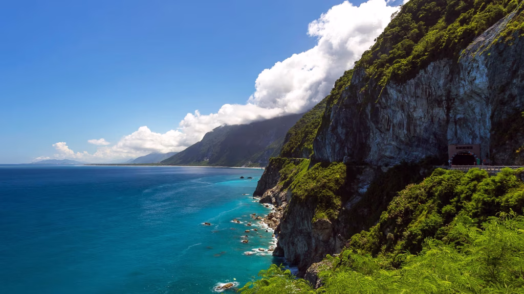 Coastal cliffside highway with a dark tunnel opening, lush greenery, and a turquoise sea under a blue sky.