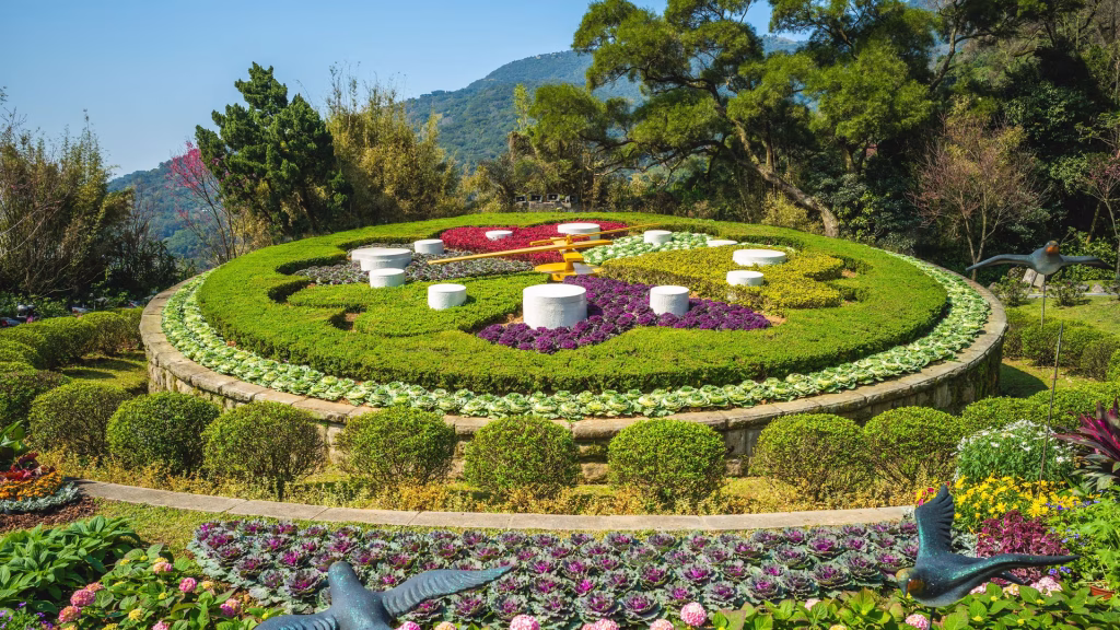 Circular terraced garden with concentric flower beds, white pedestals, and bird statues in the foreground against a blue sky and hills.