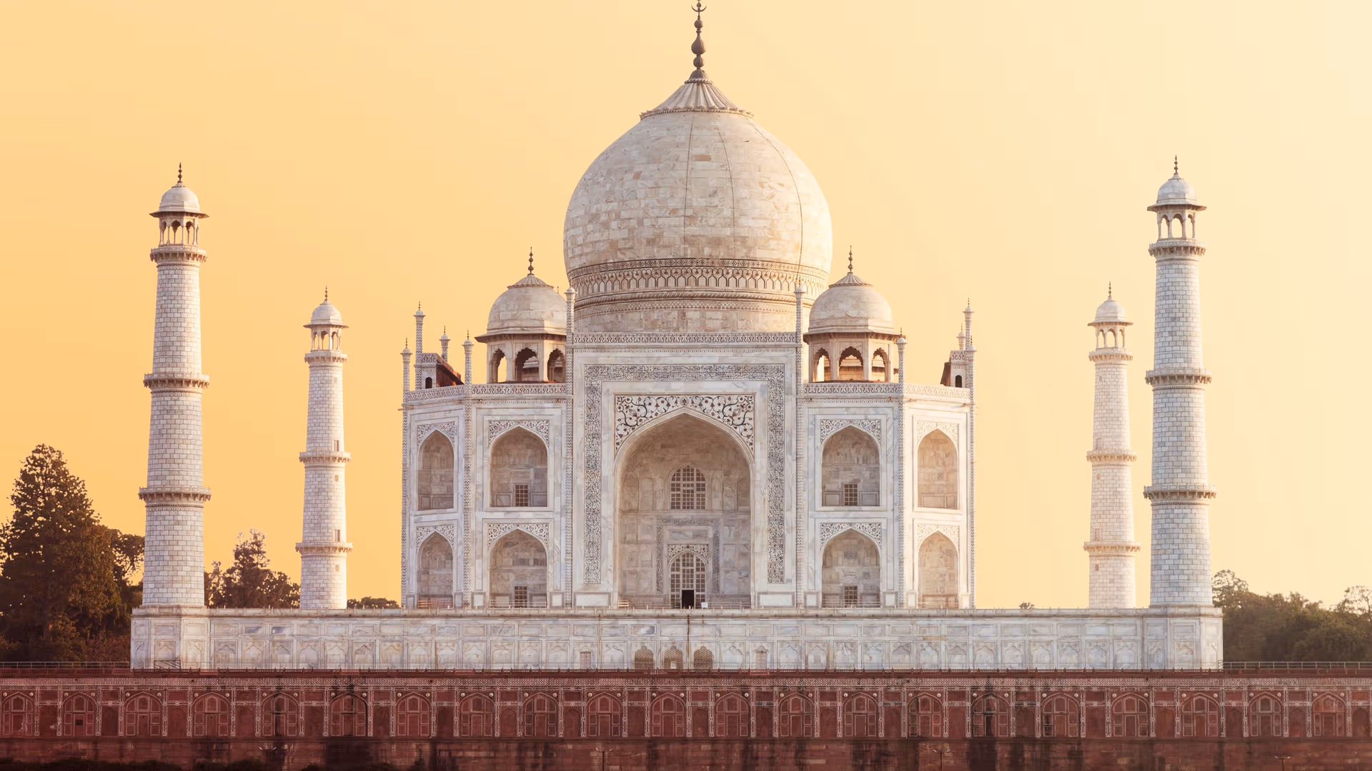 Taj Mahal mausoleum in white marble with a large central dome and four surrounding minarets against a warm orange sky at sunrise, front view.