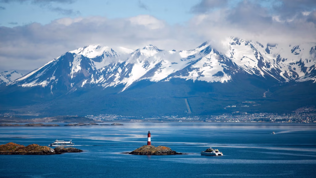 Snow-capped mountains above a calm channel with a red-and-white lighthouse and two passenger boats.