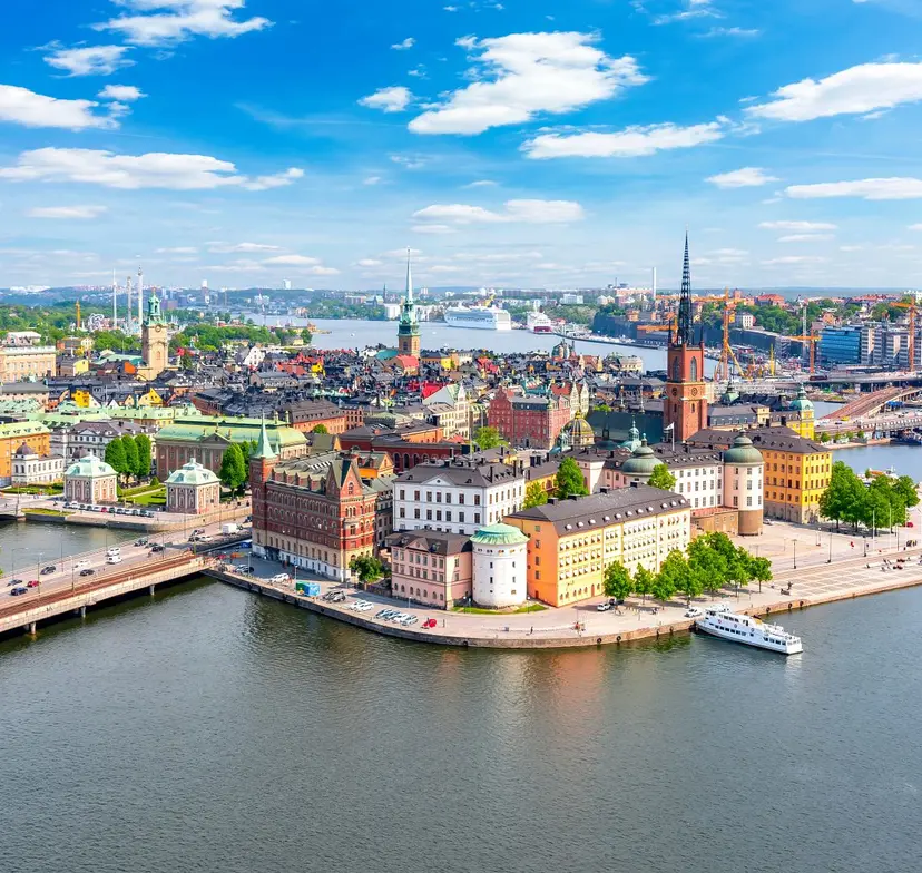 Aerial view of Stockholm's old town on a peninsula, with colorful historic buildings, a tall church spire, and a harbor under a blue sky.