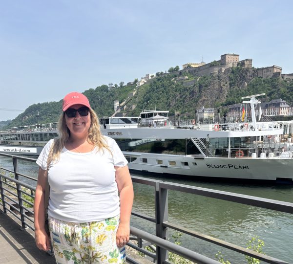 Woman in a coral cap and sunglasses stands on a riverside promenade, white top and tropical-print pants, with a large white boat named Scenic Pearl in the background and a hillside fortress beyond.