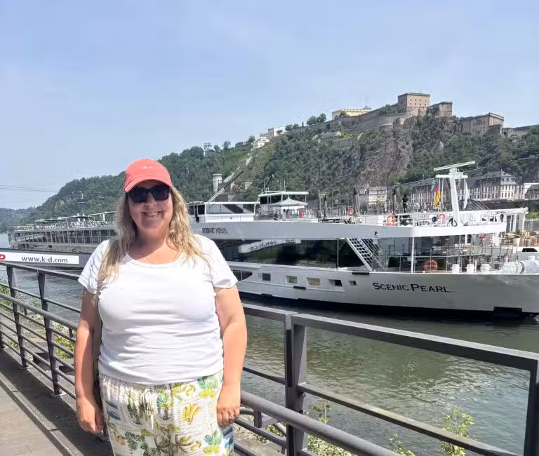 Woman in a coral cap and sunglasses stands on a riverside promenade, white top and tropical-print pants, with a large white boat named Scenic Pearl in the background and a hillside fortress beyond.