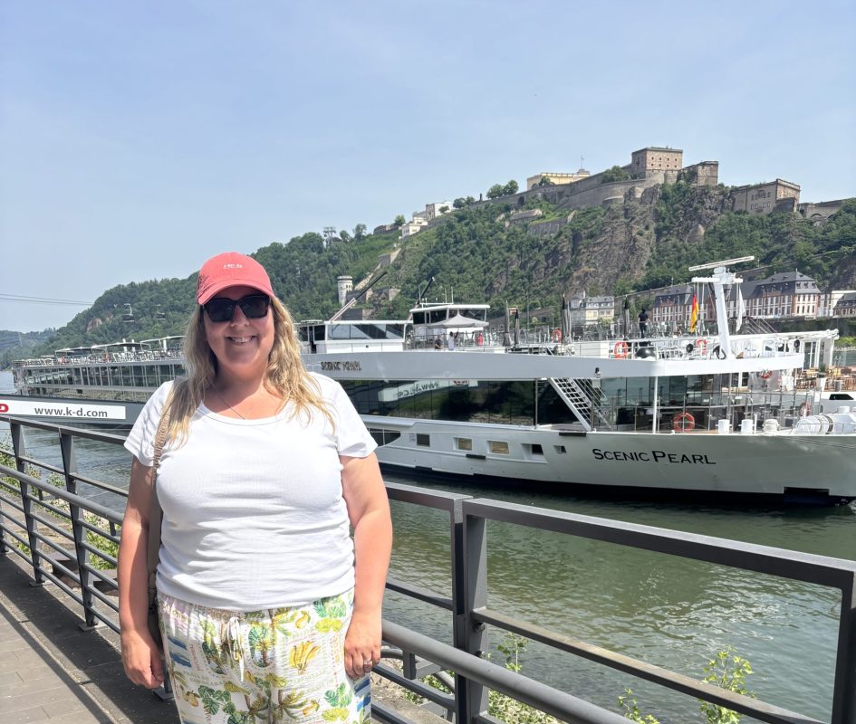 Woman in a coral cap and sunglasses stands on a riverside promenade, white top and tropical-print pants, with a large white boat named Scenic Pearl in the background and a hillside fortress beyond.