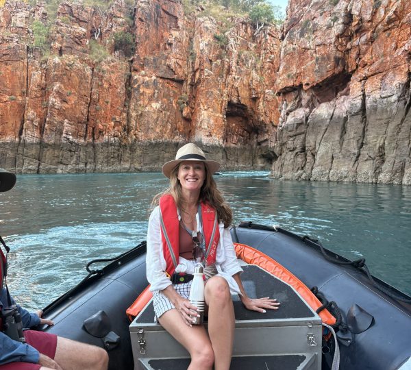 Smiling woman in a life jacket and hat sits on a small inflatable boat near red, jagged canyon walls; calm blue-green water surrounds them.]