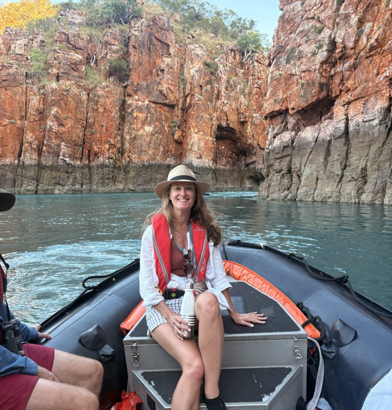 Smiling woman in a life jacket and hat sits on a small inflatable boat near red, jagged canyon walls; calm blue-green water surrounds them.]