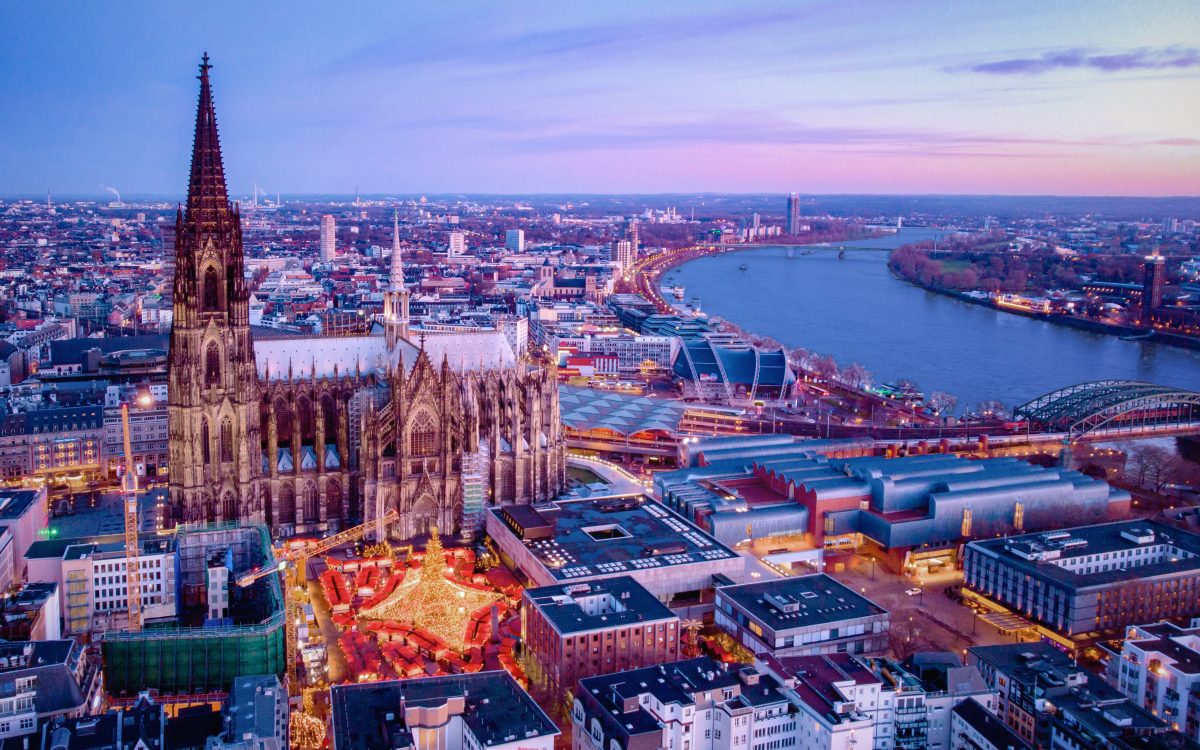 Aerial view of Cologne at dusk: the towering Cologne Cathedral with a glowing Christmas market in the foreground and the Rhine River to the right.