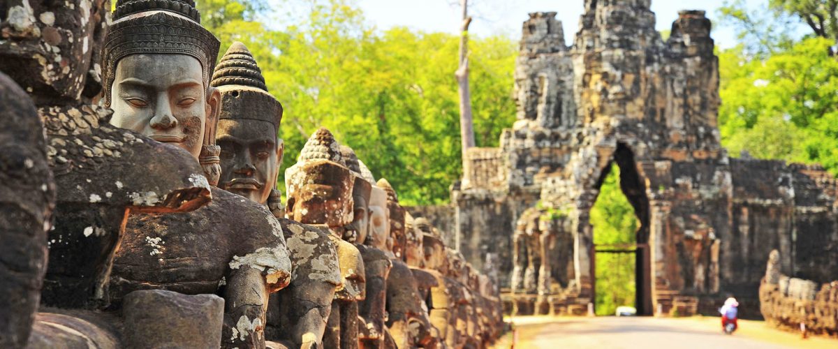 Line of weathered stone Buddha heads along a temple walkway, with a distant arch and green forest in the background.