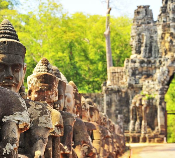 Line of weathered stone Buddha heads along a temple walkway, with a distant arch and green forest in the background.