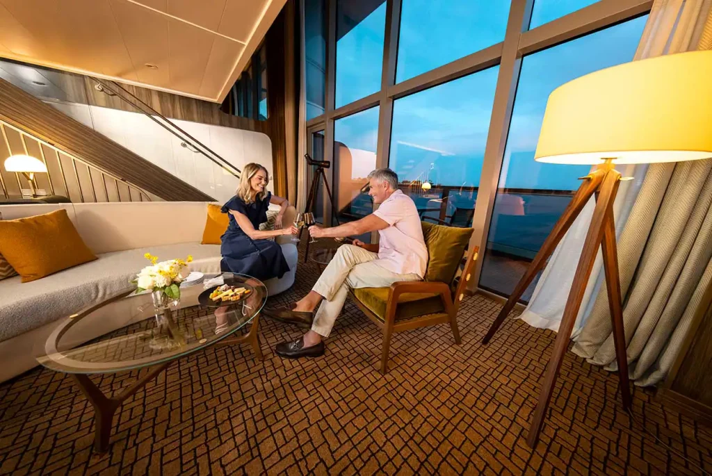Couple toasting with champagne in a modern hotel lounge by a large window overlooking blue sky, sofa with yellow cushions, and a glass coffee table.