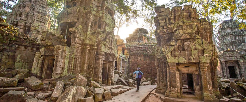 Traveler with backpack walks along a path between moss-covered ancient stone temple ruins at sunset