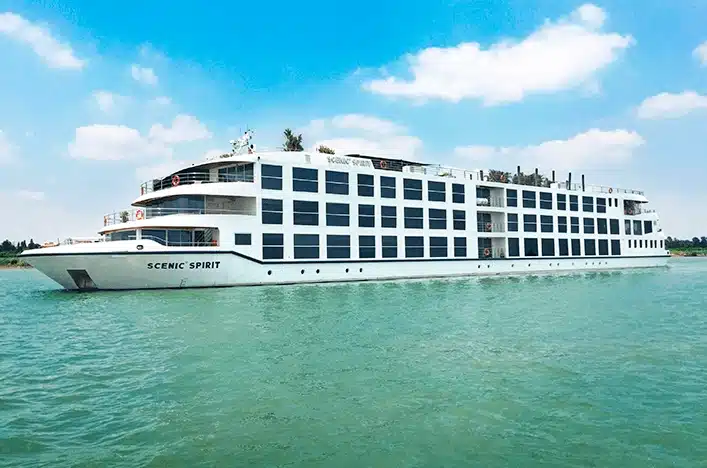 White multi-deck passenger cruise ship named Scenic Spirit sailing on turquoise water under a blue sky with scattered clouds.