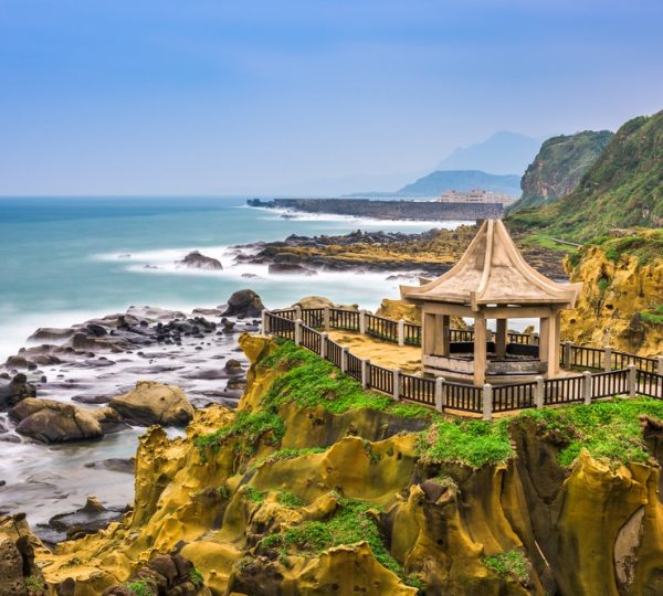 Stone pavilion atop a grassy cliff with a wooden railing overlooking rocky shoreline and blue sea far beyond.