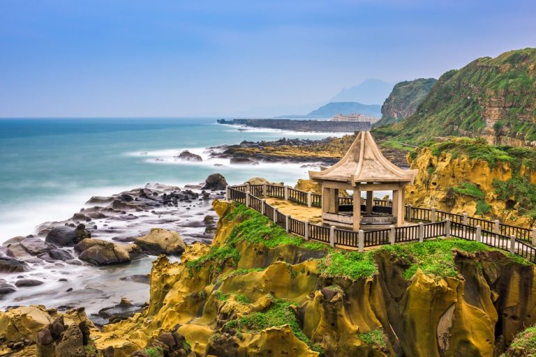 Stone pavilion atop a grassy cliff with a wooden railing overlooking rocky shoreline and blue sea far beyond.