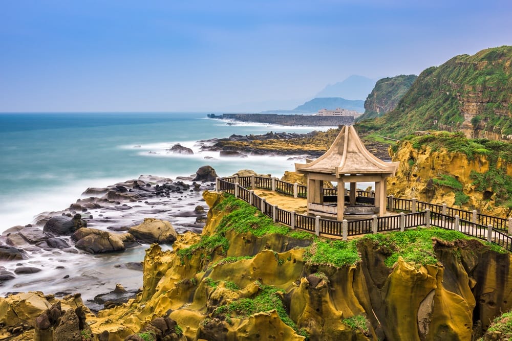Stone pavilion atop a grassy cliff with a wooden railing overlooking rocky shoreline and blue sea far beyond.