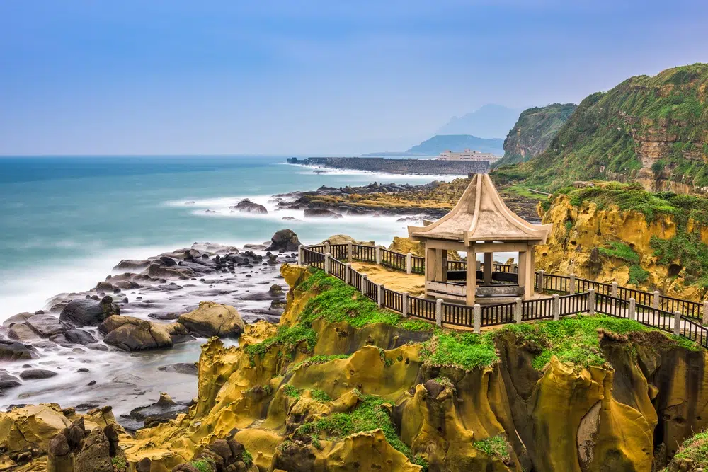 Stone pavilion atop a grassy cliff with a wooden railing overlooking rocky shoreline and blue sea far beyond.