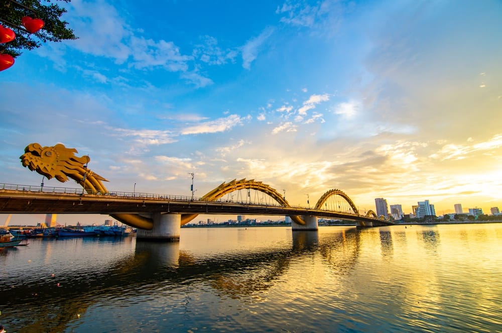 Dragon-headed arch bridge spanning a calm river at sunset, with city buildings in the background and reflections on the water.