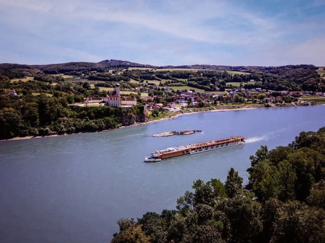 Aerial view of a riverside town with a white church, green hills, and a long tour boat on calm water.