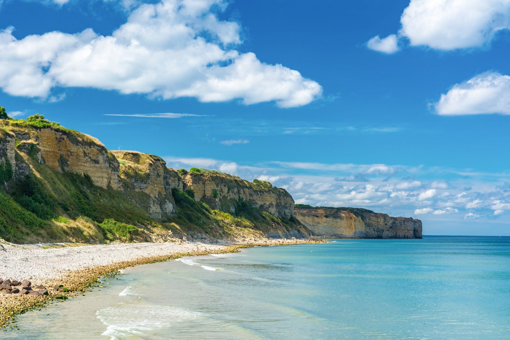Cliff coastline with green vegetation, a pebbled beach, and turquoise sea under a bright blue sky with white clouds.