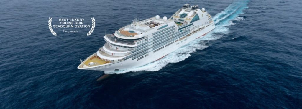 White luxury cruise ship sailing through deep blue ocean, wake streaming behind it, with an award badge on the left reading 'Best Luxury Cruise Ship Seabourn Ovation'.