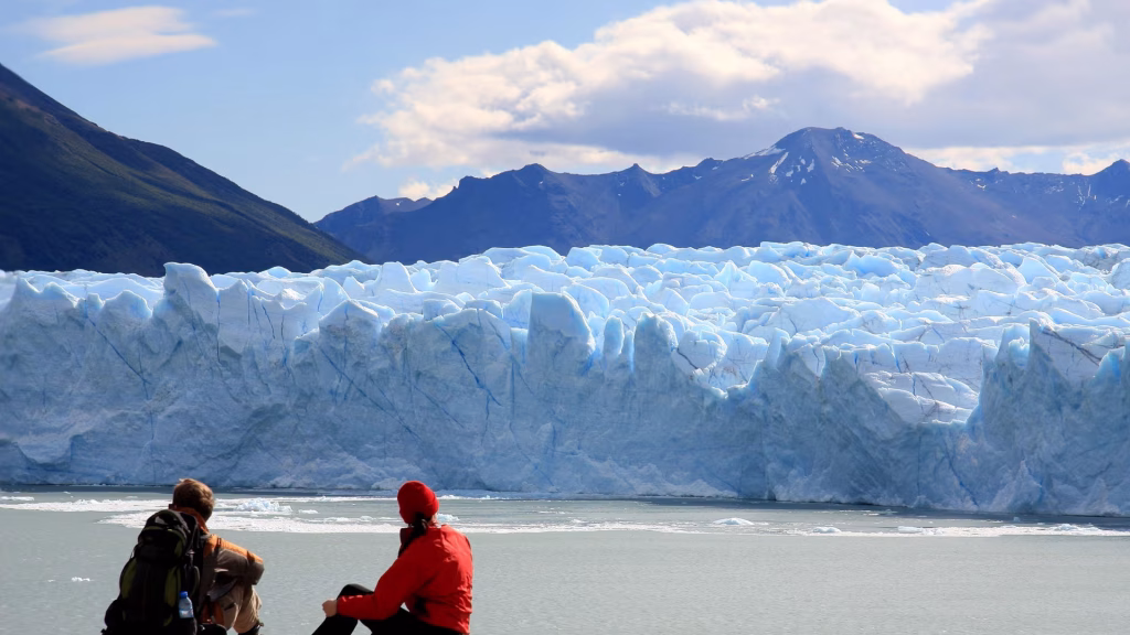Two hikers in a red jacket and backpack sitting before a towering blue glacier face.